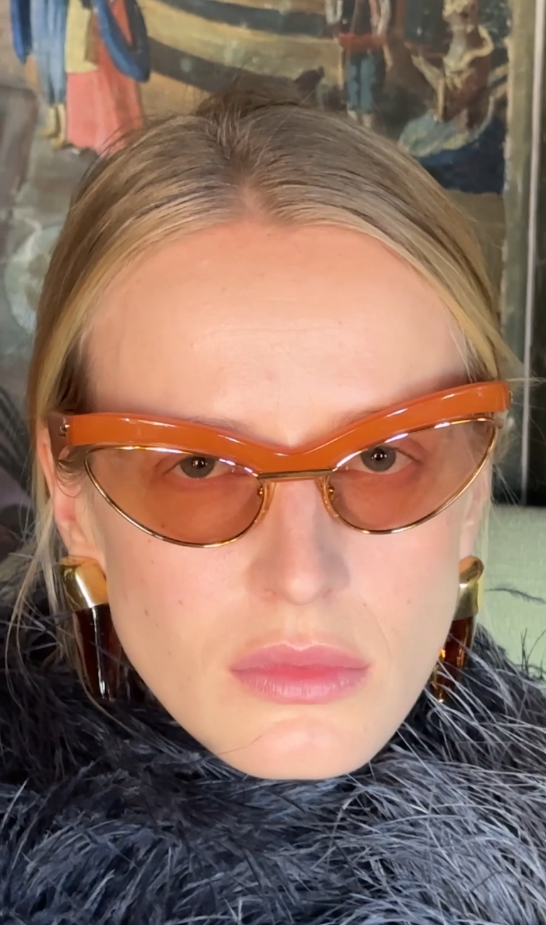 Model in orange cat-eye Bottega Veneta sunglasses with a feathered collar (still from the Summer 2026 film)