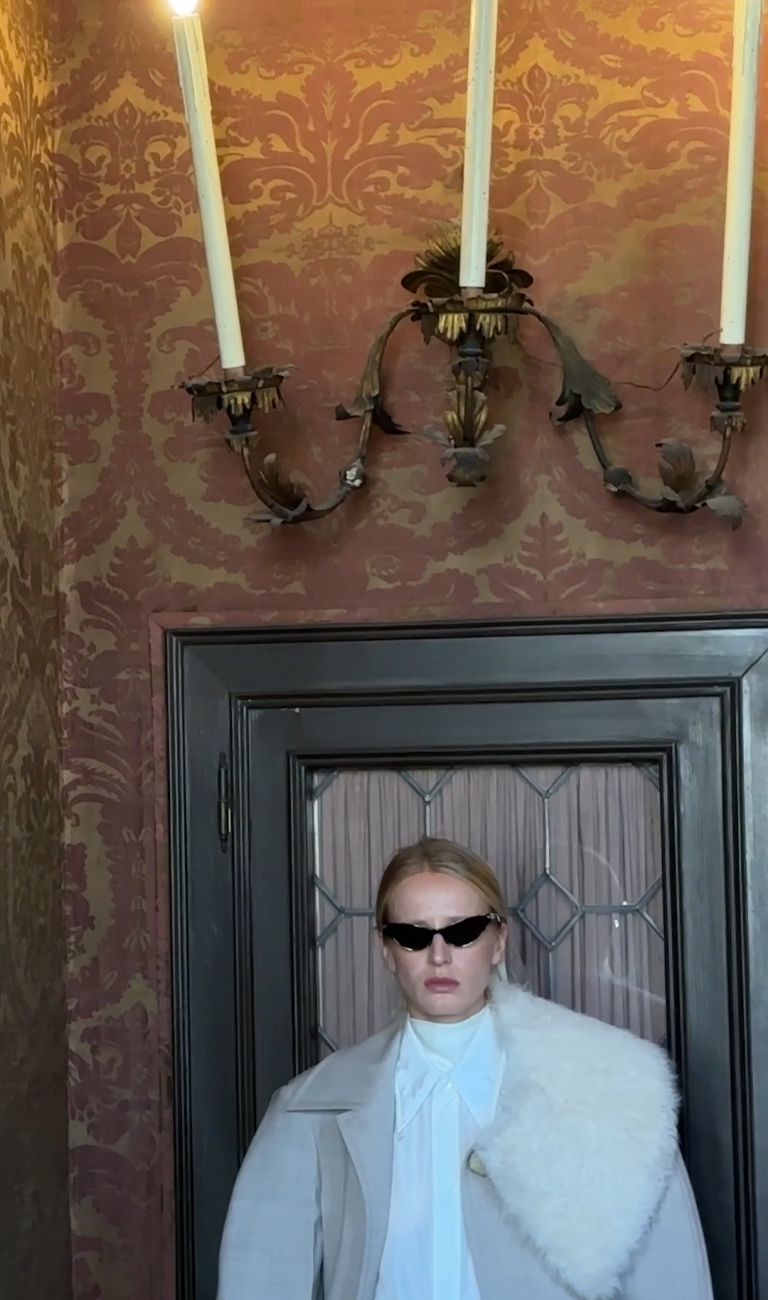 Model in black Bottega Veneta sunglasses standing in a doorway under candles (still from the Summer 2026 film)
